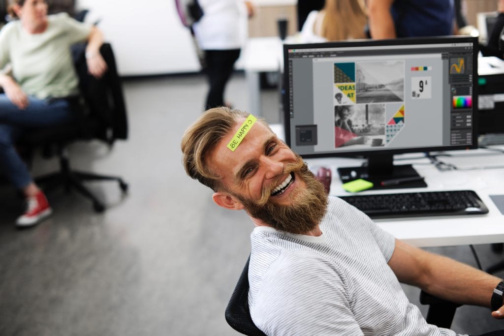 Man in an office laughing with a sticky note saying 'Be happy' stuck to his forehead.