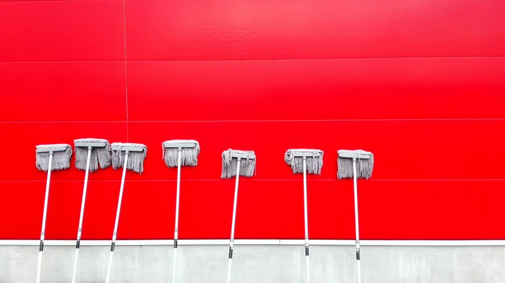 Seven mops leant against a red wall.