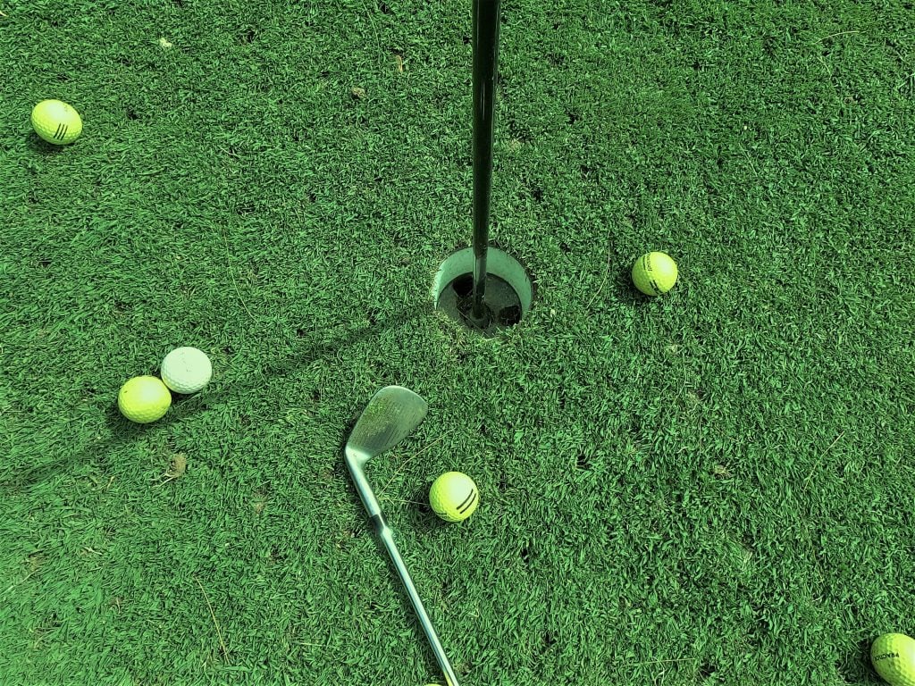 Six golf balls and a golf club around a hole on a putting green. 