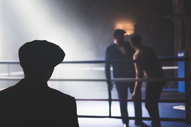 Two men in a boxing ring while a man in a hat watches from the shadows in the foreground.