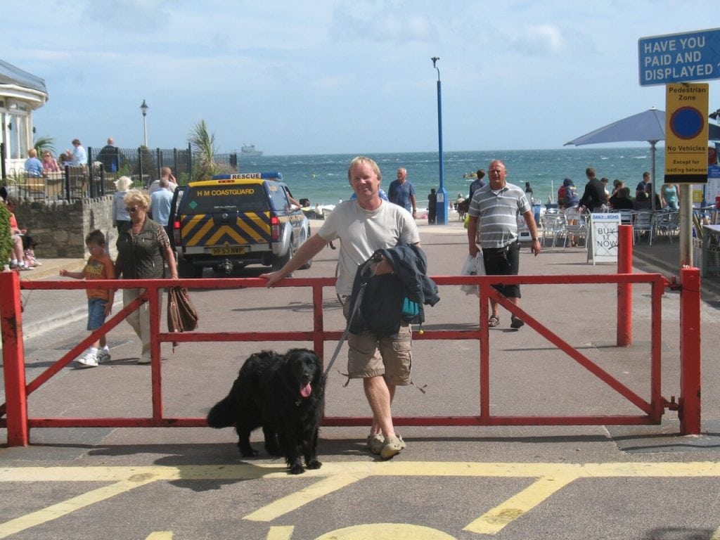 Chris Croft with his dog, standing at a gate with the beach and sea in the background.