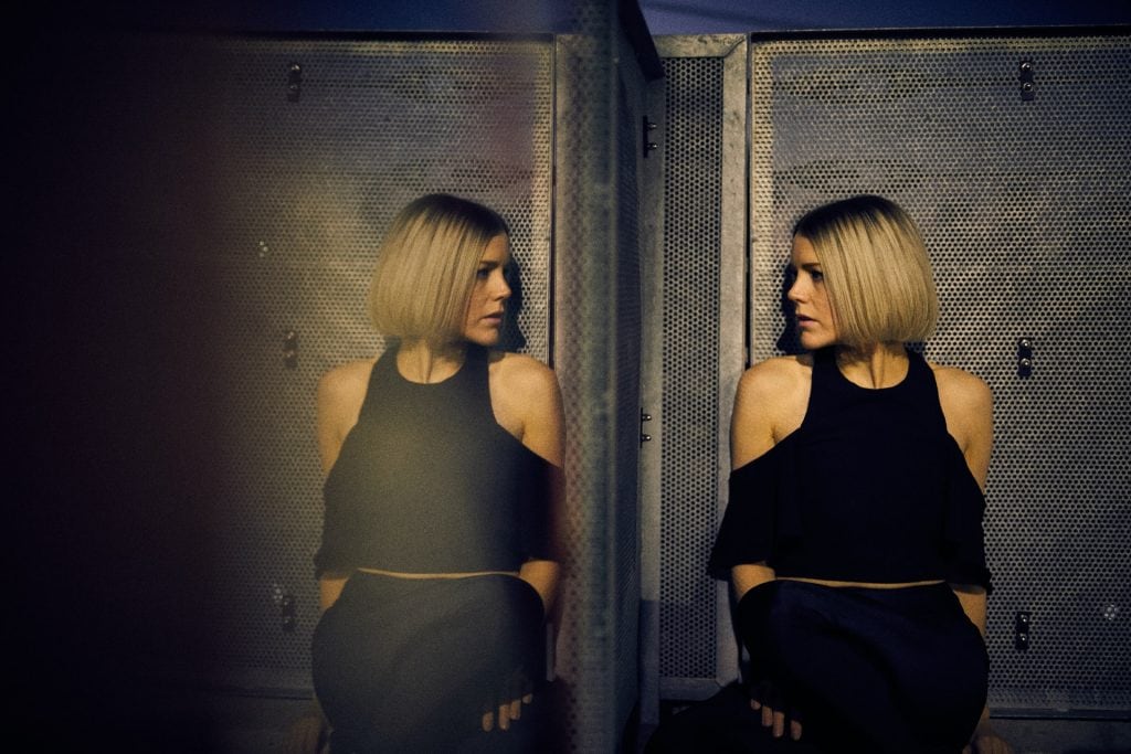 Blonde woman leaning against a wall, looking into a large mirror beside her.