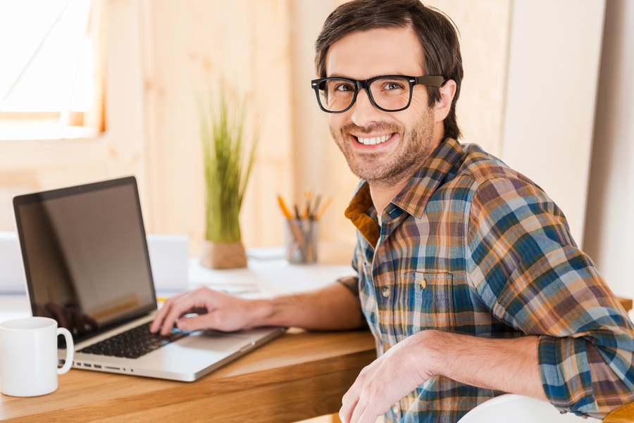 young man in glasses working on his laptop and looking at camera over shoulders while sitting at his working place