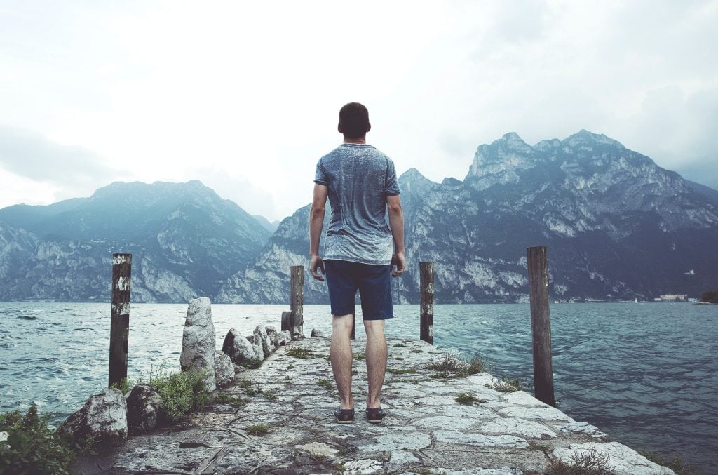 back view of man standing on rocky pier overlooking lake and mountains